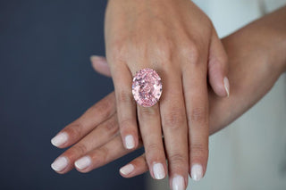 Close-up of a hand wearing a large oval-cut pink diamond ring, displayed against a dark background with neatly manicured nails.