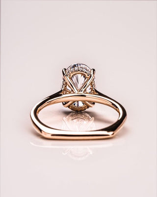 The ring showcases a hidden halo of small diamonds around the base of the center stone and a subtly squared band, resting on a glossy, reflective surface that highlights the intricate craftsmanship and brilliance of the diamond.