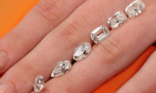 Close-up of different diamond shapes — oval, pear, marquise, emerald, and cushion cuts — displayed on fingers against an orange background.