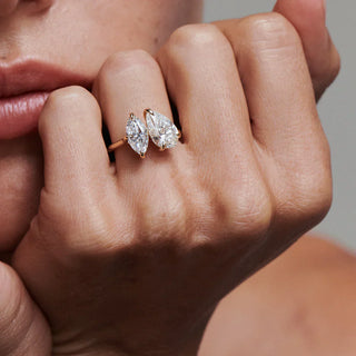 Close-up of a hand wearing a toi et moi diamond ring featuring two pear-shaped diamonds set in opposite directions on a thin rose gold band, styled near the lips for a bold and elegant look.