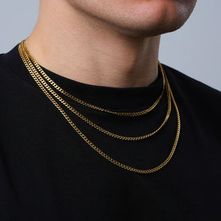 Close-up of a man wearing three layered gold Cuban chains over a black t-shirt, showcasing different thicknesses and lengths of the chains.