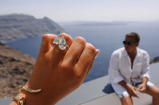 Toi et Moi engagement ring with pear and emerald cut diamonds on hand overlooking Santorini sea view