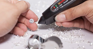 Handheld diamond tester being used to check the authenticity of a loose round diamond surrounded by scattered stones and a jeweler's loupe