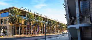 Exterior view of the Gold and Diamond Park in Dubai featuring palm lined walkways, retail storefronts and the main building façade under clear blue skies.