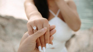 A custom engagement ring in a velvet box held by a hand, ready for a proposal, with a blurred romantic setting in the background.