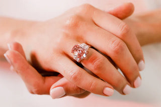 **Alt Image Text:** Close-up of a hand wearing a large cushion-cut pink diamond ring flanked by tapered baguette diamonds, set in a delicate prong setting, styled with soft pink manicure.