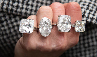 Close-up of a hand adorned with multiple large diamond rings in different cuts, including oval, marquise, emerald, and princess, all set in platinum.