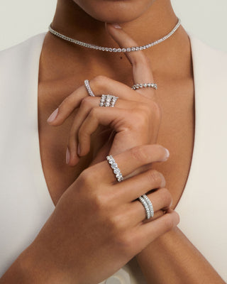     Close up of a woman wearing a diamond tennis necklace and several sparkling diamond rings, styled with a white sleeveless outfit.