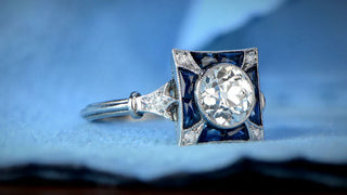 Vintage-inspired diamond ring with a round brilliant center stone framed by blue sapphire accents and intricate detailing, placed on a soft blue fabric background.