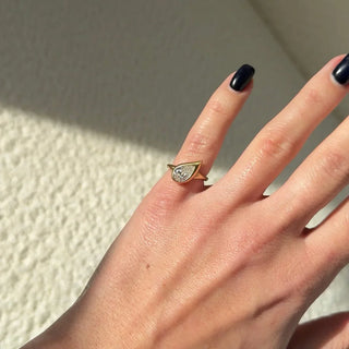 Daylight shot of a hand with a pear-cut bezel-set diamond pinky ring in yellow gold, captured in natural light with a soft shadow on a textured cream wall.
